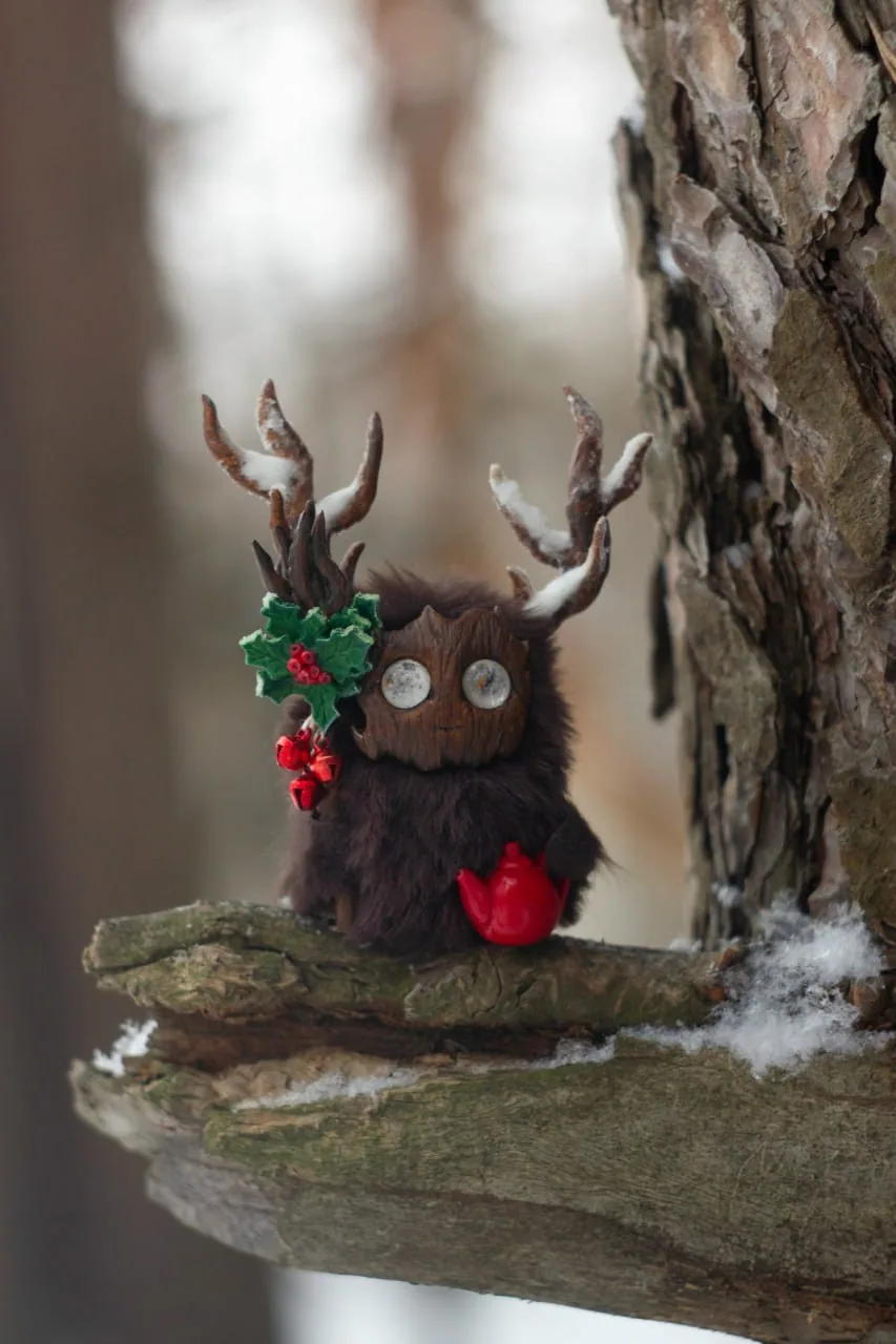 Holly Spirit — handmade chocolate-brown wool Fluffy creature with snow-dusted branching antlers, a holly-and-red-berries decoration with jingle bells, and a tiny red teapot, sitting on a snowy log | Gnomenlands